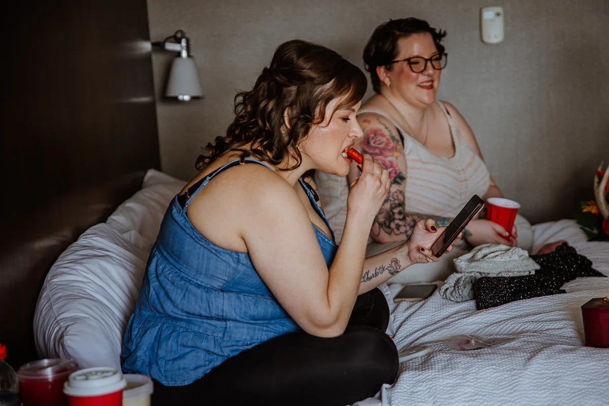 Two friends getting ready on the bed before the ceremony, candid moment applying lipstick