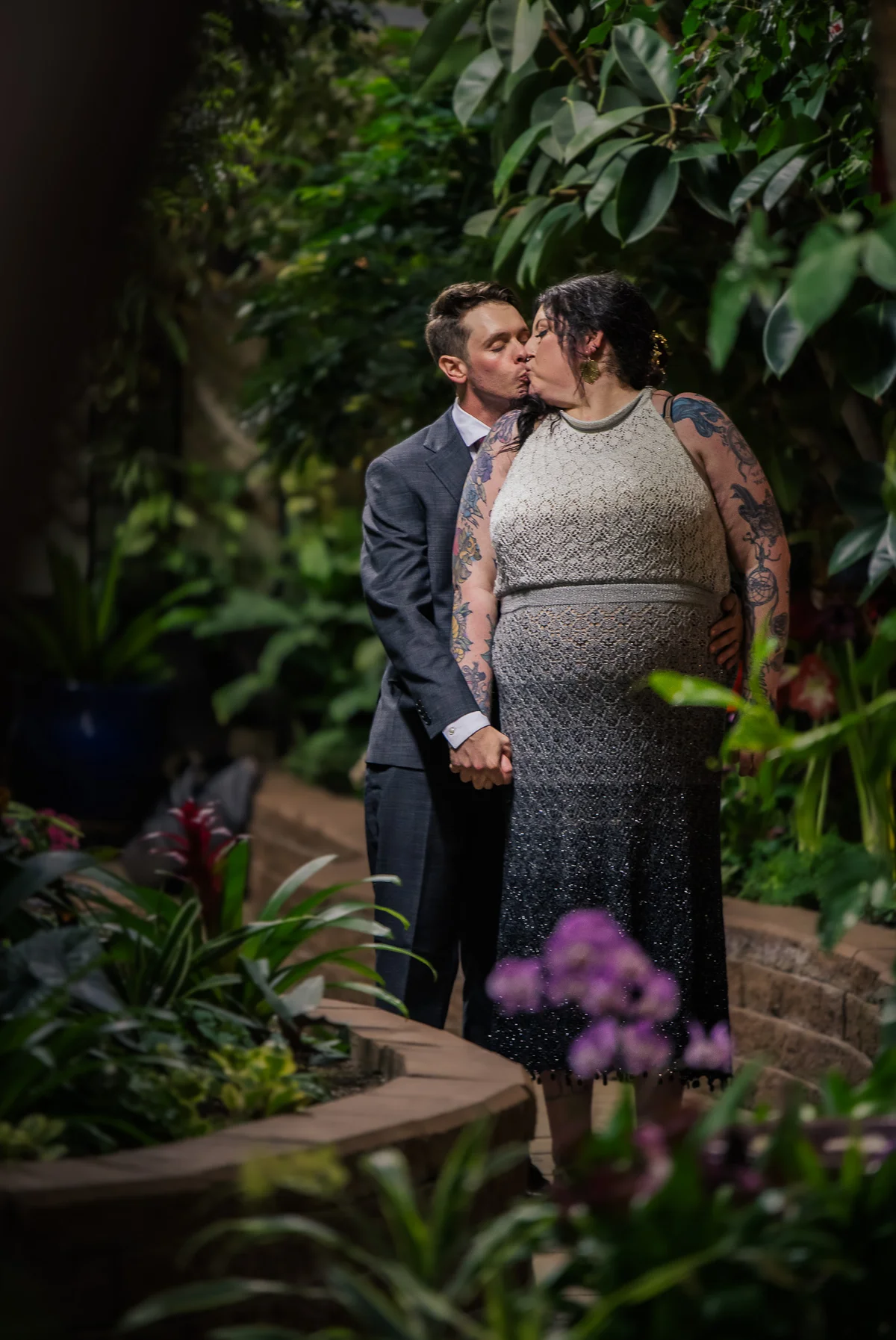 Couple kissing inside a lush greenhouse, orchids blooming in the foreground