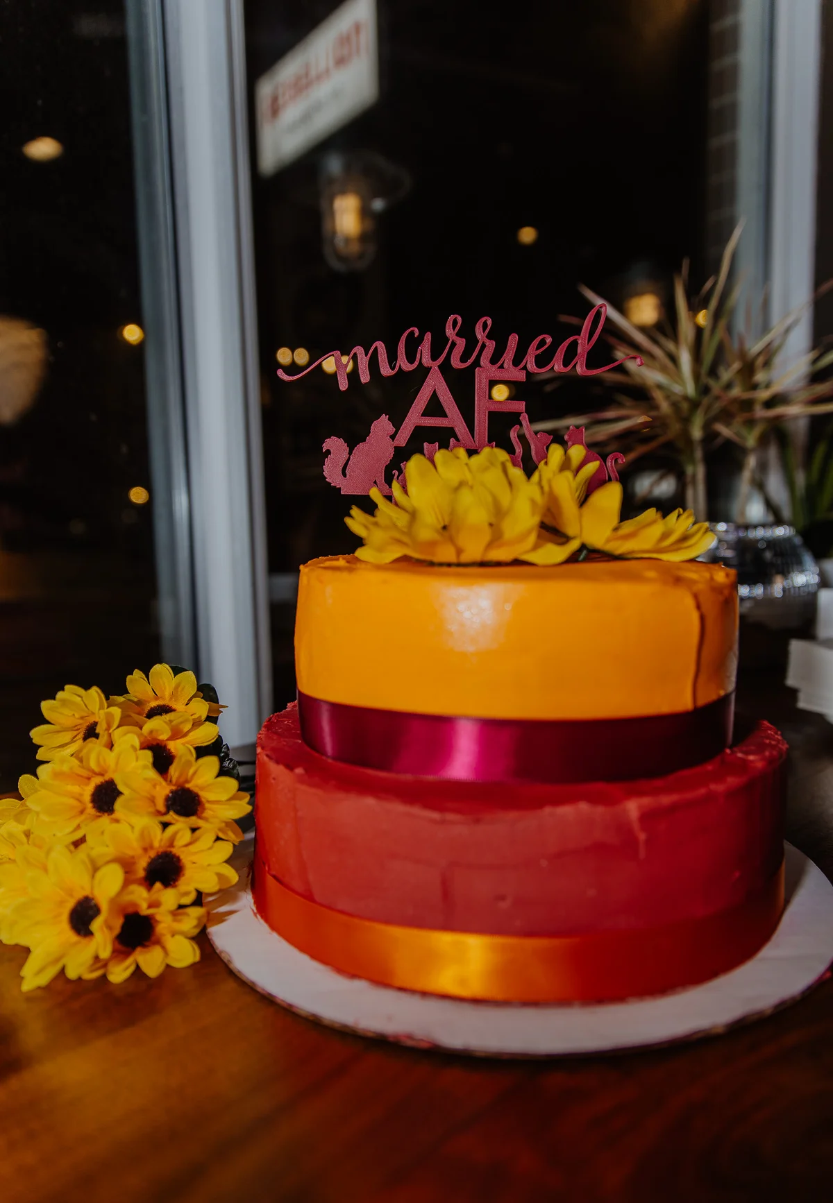 Two-tier red and orange wedding cake with a 'married AF' topper, bright sunflowers at its base