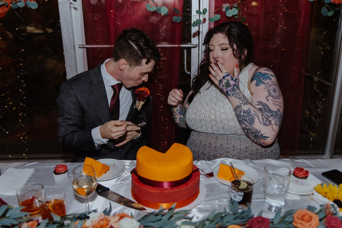 Bride and groom sharing the first bite of cake at their reception, tattooed and laughing