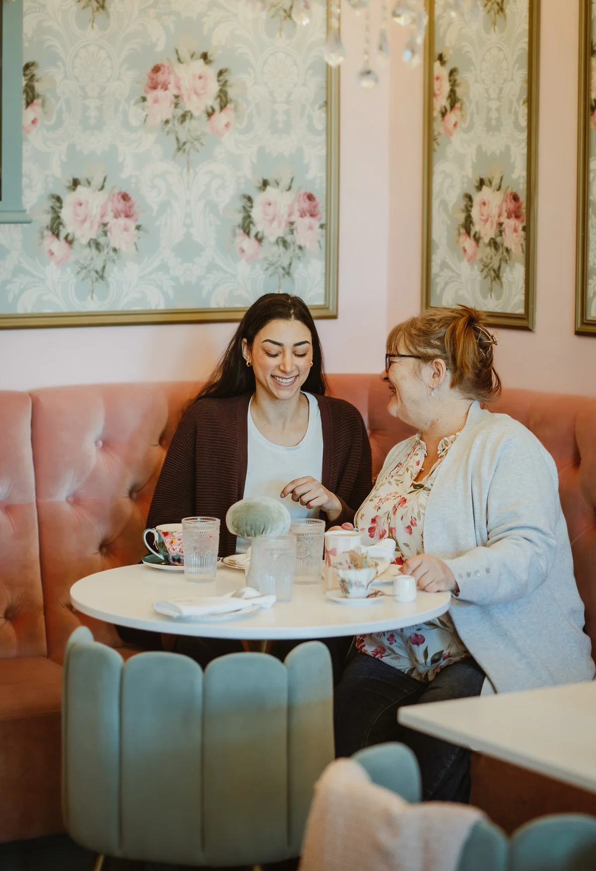 Two women laughing together over afternoon tea in a floral-wallpapered booth
