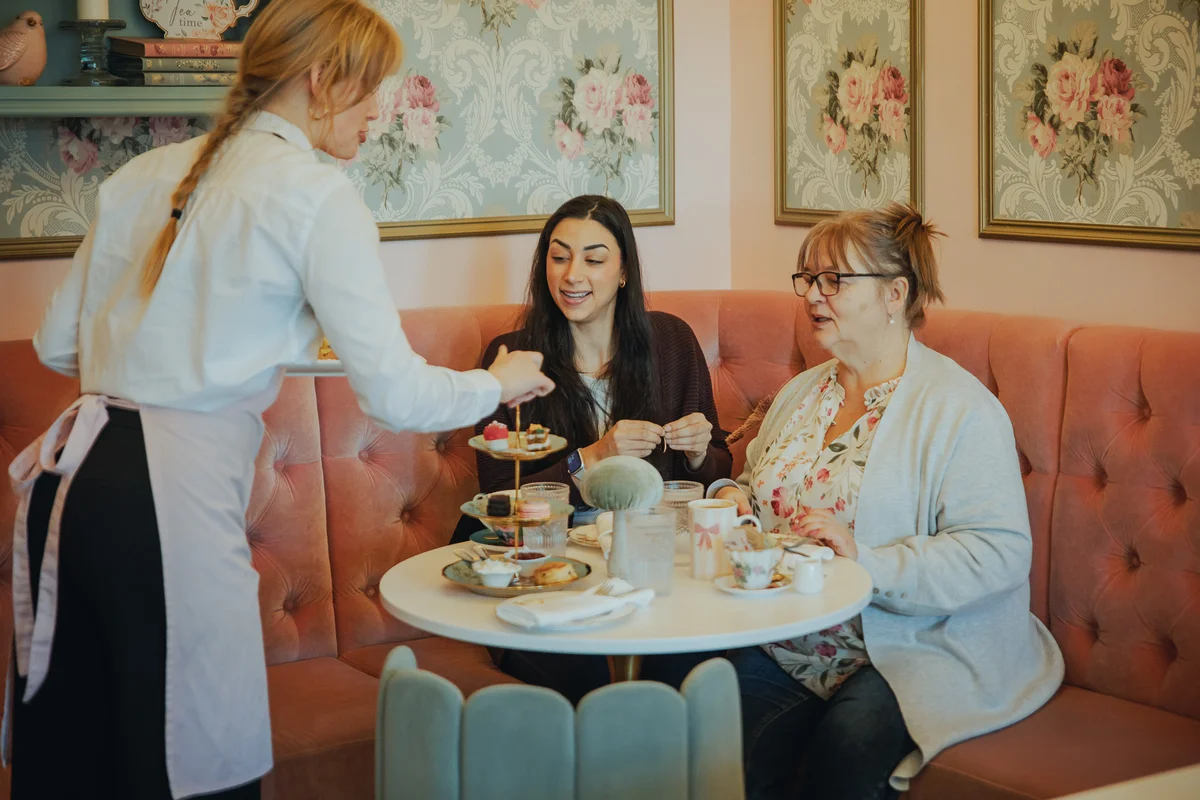 Waitress serving a tower of tea-time desserts to two guests seated in a pink booth