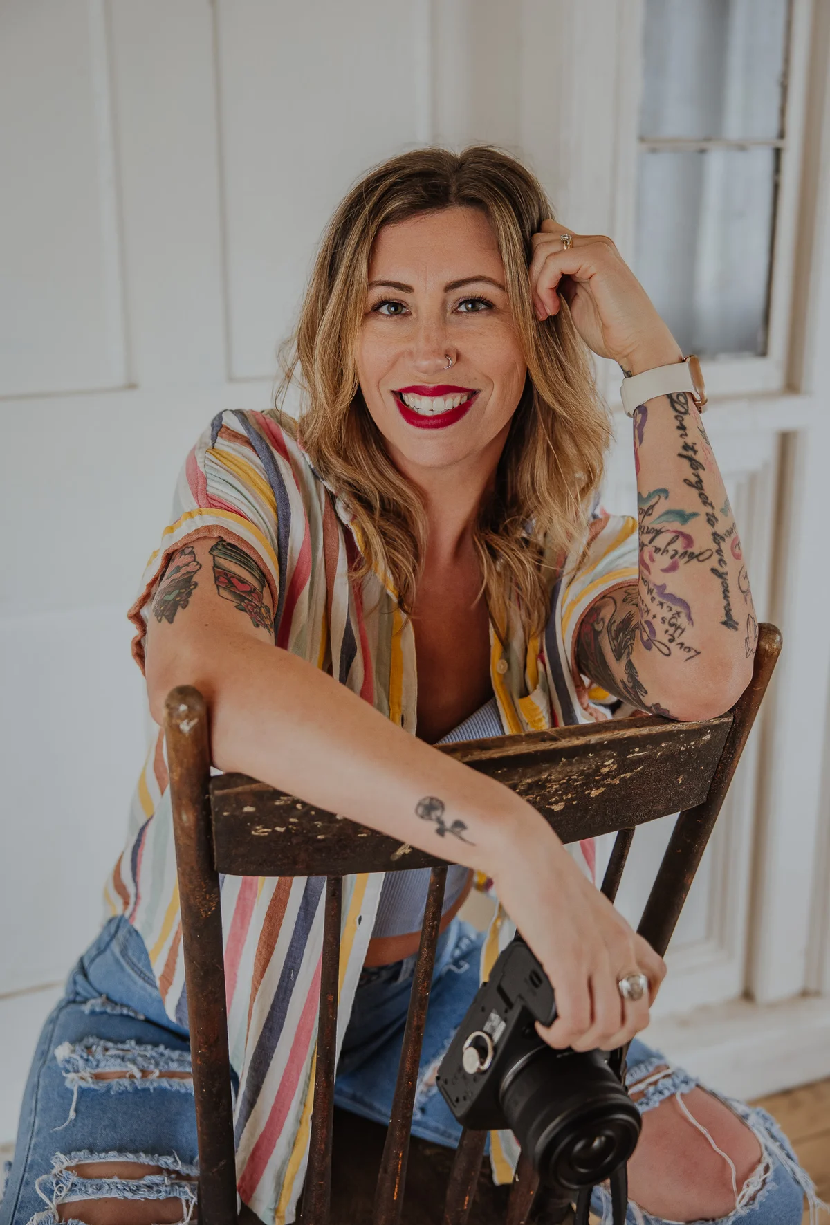 Karlene sitting backwards on a vintage wooden chair, smiling with camera in hand